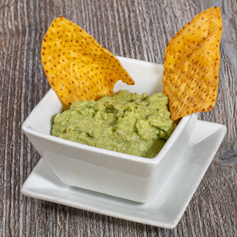 A white square bowl of guacamole with two tortilla chips on a rustic wooden surface.