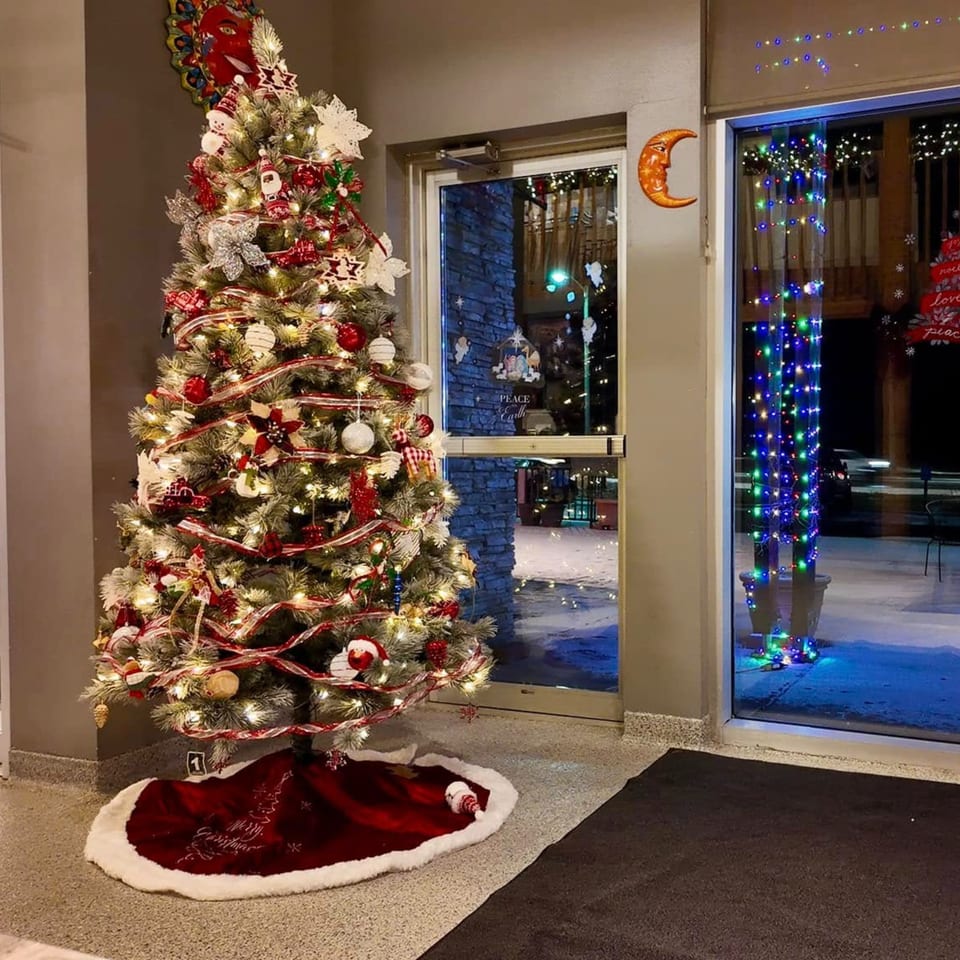 A festive Christmas tree with red and white decorations stands beside a glass door with colorful lights.